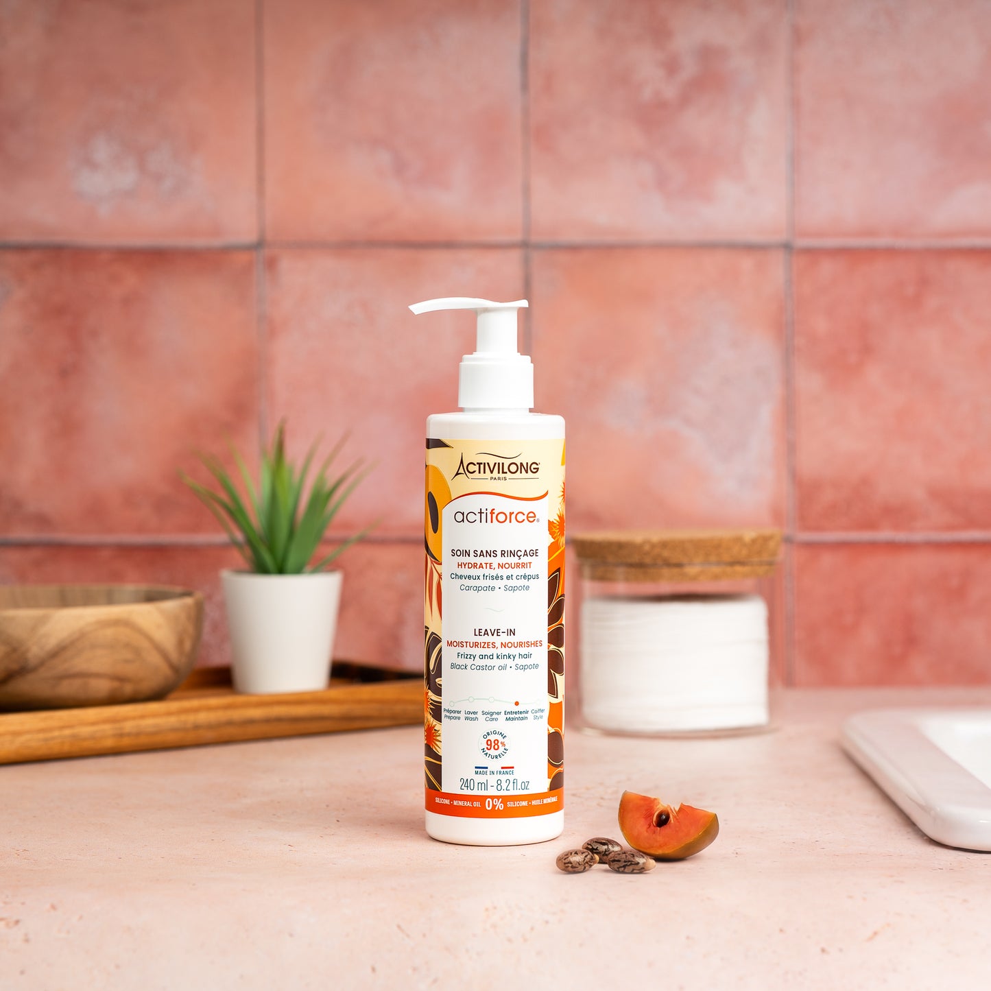 Visuel d'un Flacon de 240 ml du Soin Sans Rinçage Actiforce sur un plan de salle de bain terracotta, avec graines de carapate et tranche de sapote en arrière plan