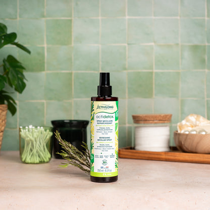 Visuel d'un Flacon du Spray Micellaire Rafraîchissant Actidetox sur un plan de salle de bain vert, avec brin de romarin et accessoires en arrière plan