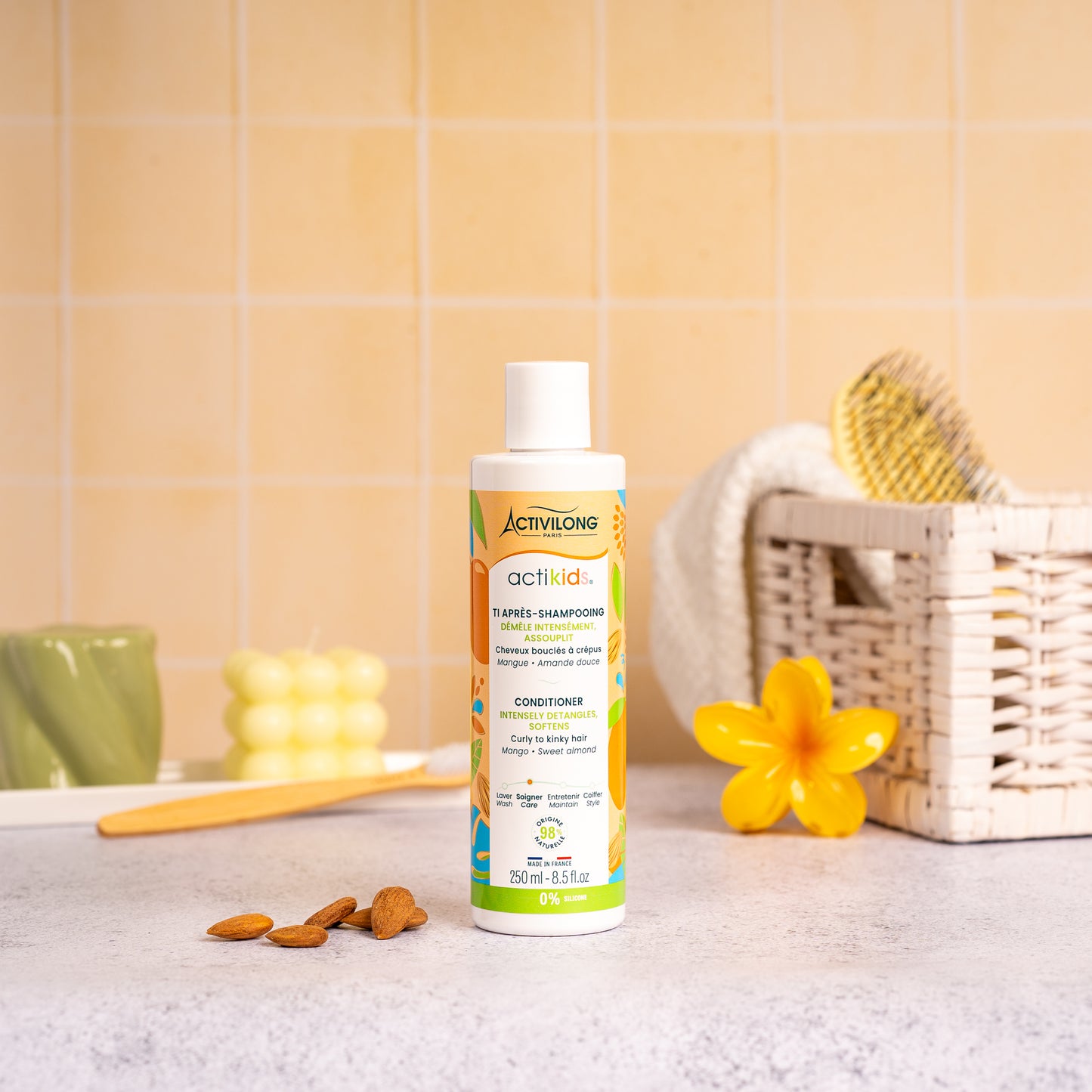 Visuel d'un Flacon de 250 ml du Ti Après-Shampooing Actikids sur un plan de salle de bain jaune, avec une fleur de frangipanier, un panier en osier garni d'une brosse et des amandes en arrière plan