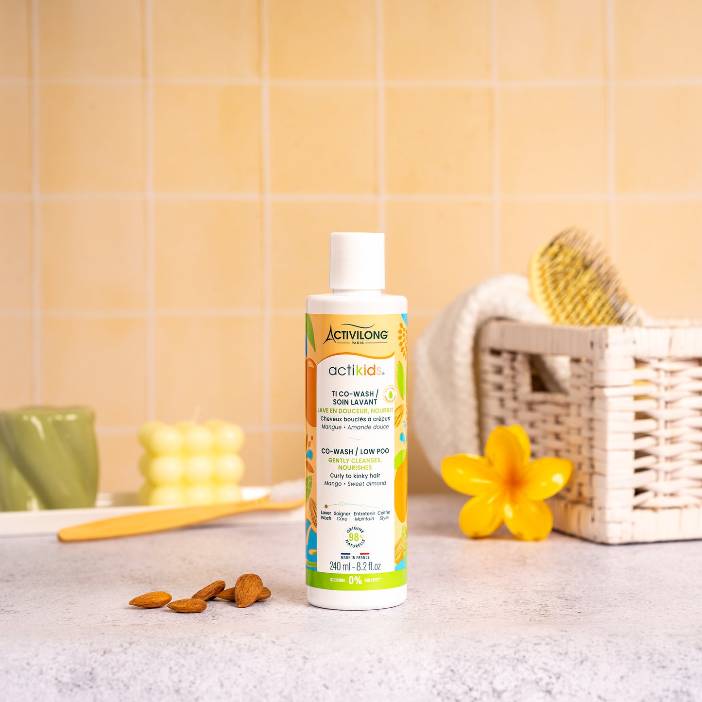 Visuel d'un Flacon du Ti Co-Wash Actikids sur un plan de salle de bain jaune, avec une fleur de frangipanier, un panier en osier garni d'une brosse et des amandes en arrière plan. 