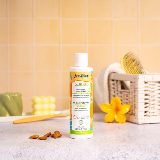 Visuel d'un Flacon du Ti Co-Wash Actikids sur un plan de salle de bain jaune, avec une fleur de frangipanier, un panier en osier garni d'une brosse et des amandes en arrière plan. 