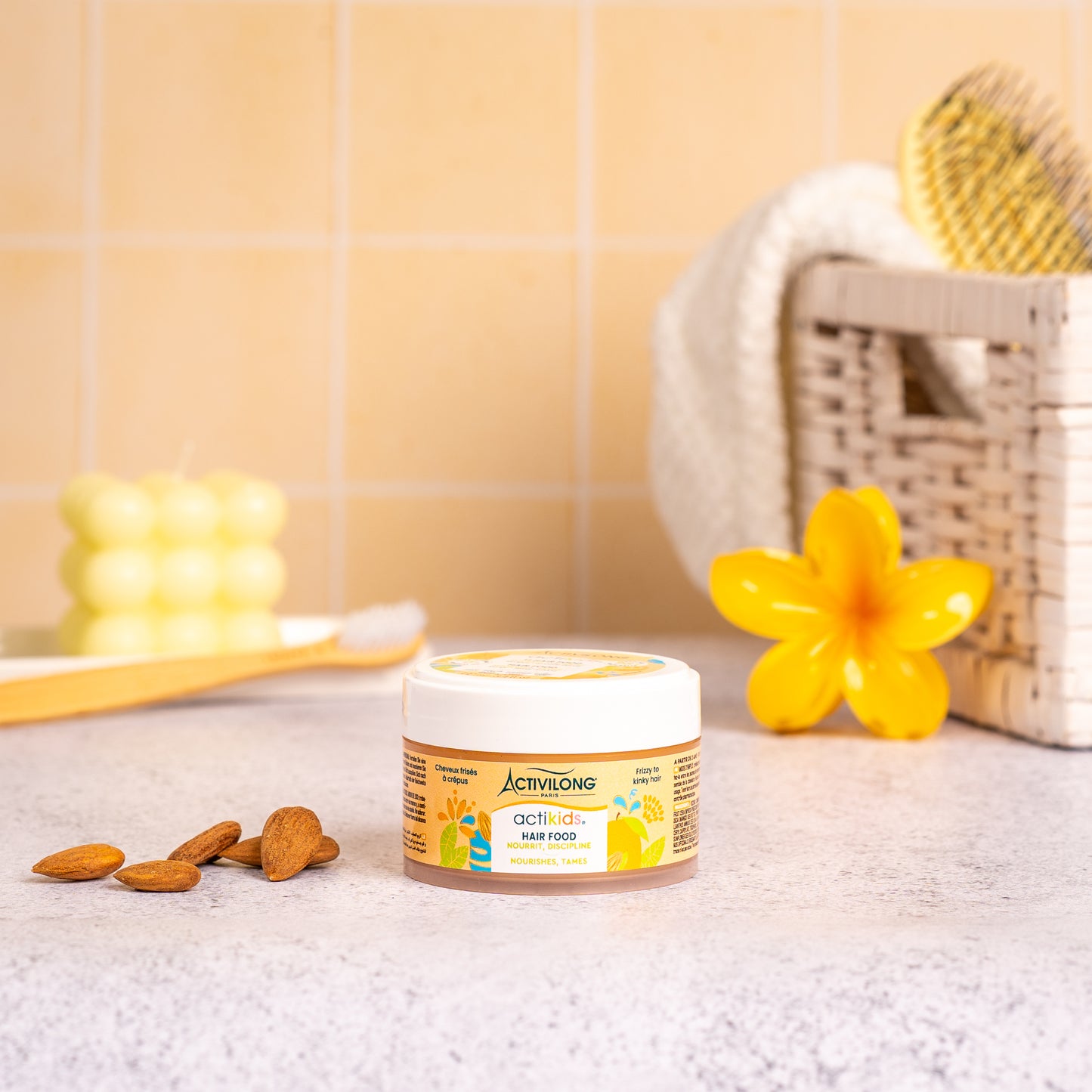 Visuel d'un Pot de 100 ml du Hair Food Actikids sur un plan de salle de bain jaune, avec brosse, fleur jaune et amandes en arrière plan