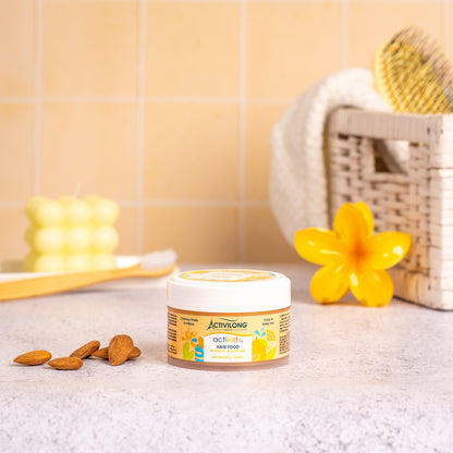 Visuel d'un Pot de 100 ml du Hair Food Actikids sur un plan de salle de bain jaune, avec brosse, fleur jaune et amandes en arrière plan
