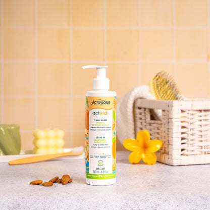 Visuel Lifestyle d'un flacon de 240 ml du Ti Milkshake / Leave-In Actikids posé sur un plan de salle de bain jaune, avec une fleur de frangipanier, un panier en osier garni d'une brosse et des amandes en arrière-plan.