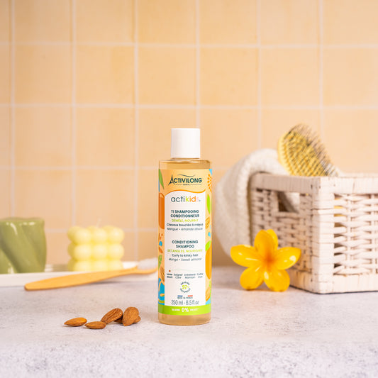 Flacon de 250 ml du Ti Shampooing Conditionneur Actikids posé sur un plan de salle de bain jaune, avec une fleur de frangipanier, un panier en osier garni d'une brosse et des amandes en arrière-plan.