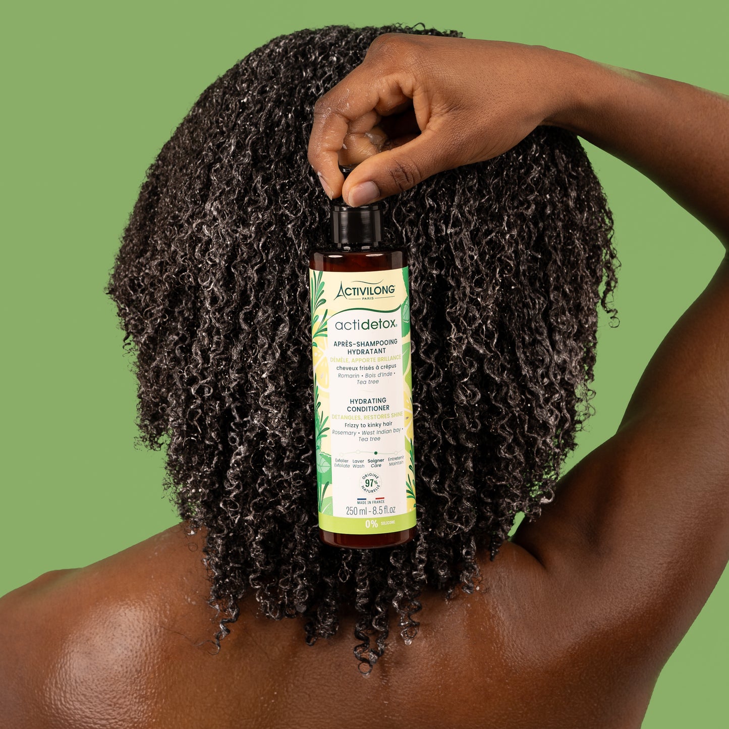 Visuel de dos d'une femme aux cheveux crépus tenant le flacon Après-shampooing hydratant Actidetox Activilong devant sa chevelure, sur fond vert