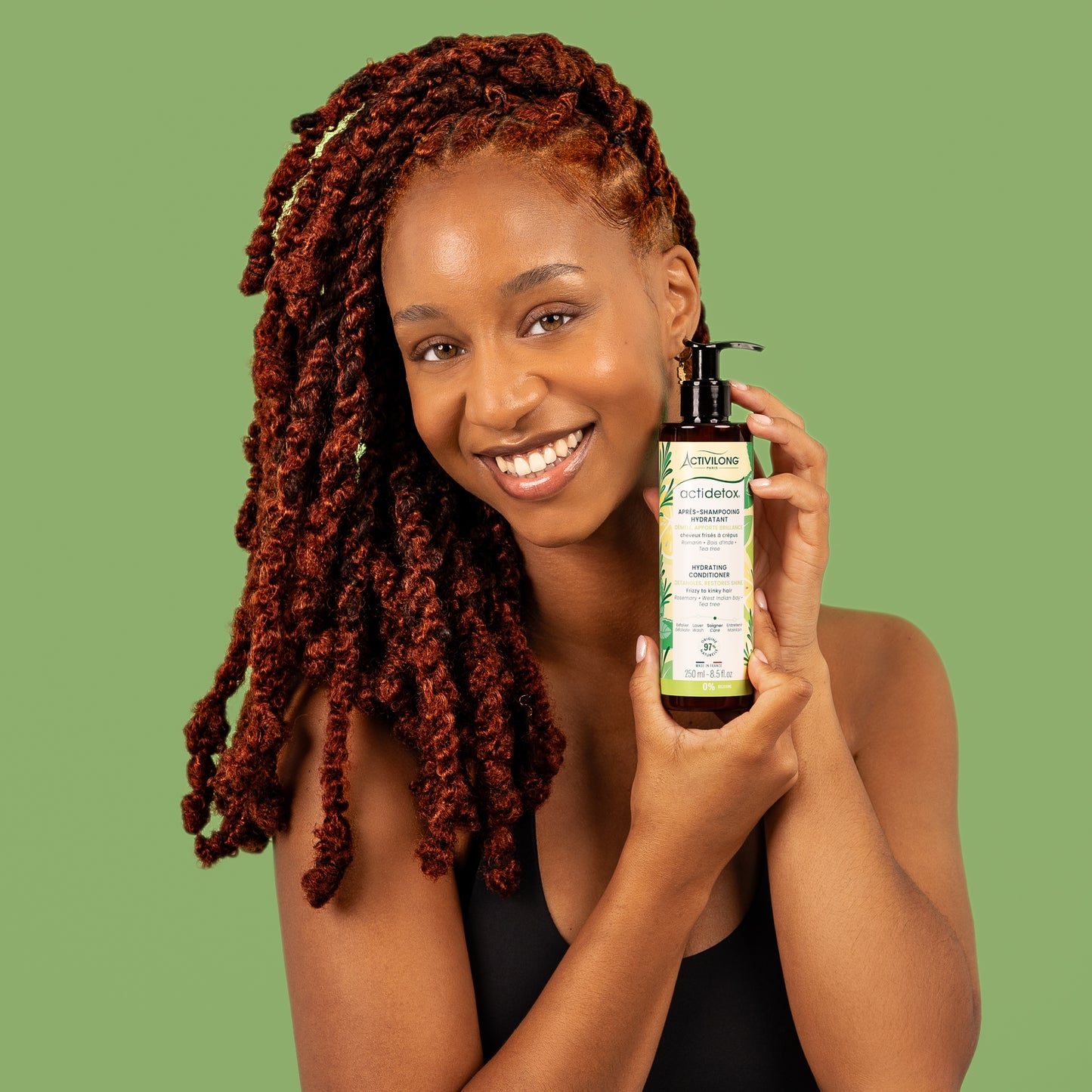 Visuel d'une femme souriante aux longues locks auburn tenant le flacon Après-shampooing hydratant Actidetox Activilong près de son visage, sur fond vert