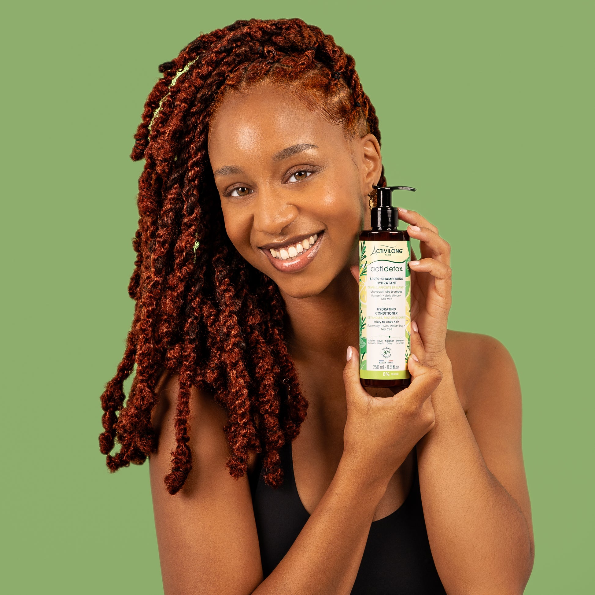 Visuel d'une femme souriante aux longues locks auburn tenant le flacon Après-shampooing hydratant Actidetox Activilong près de son visage, sur fond vert