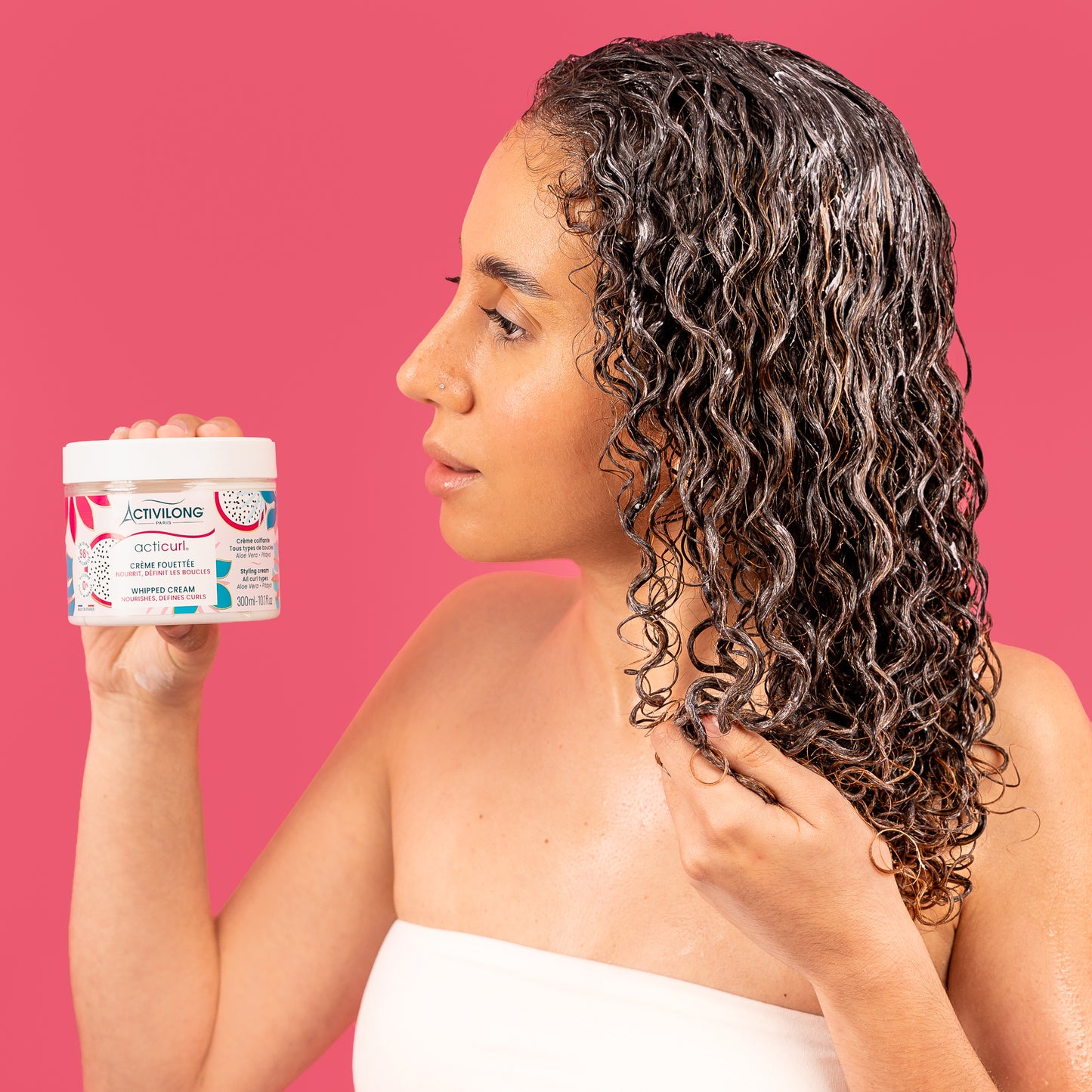 Visuel d'une femme de profil aux longues boucles définies tenant le pot Crème fouettée Acticurl Activilong et travaillant une mèche, sur fond rose vif