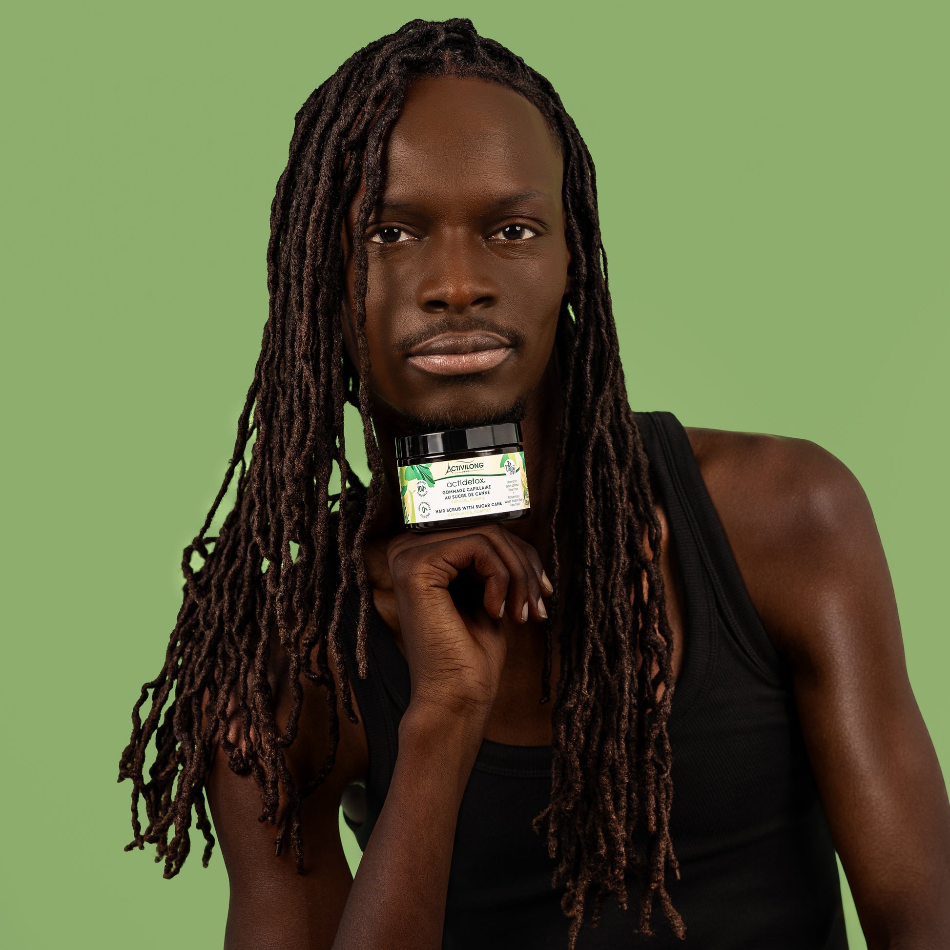 Visuel d'un homme aux longues locks tenant le pot Gommage capillaire au sucre de canne Actidetox Activilong sous son menton, sur fond vert