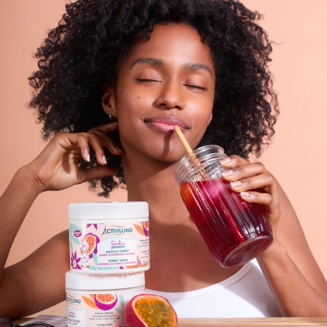 Visuel d'une femme aux cheveux bouclés sirotant une boisson rose à la paille, avec les pots Masque sorbet et Crème onctueuse Tendre Passion au premier plan