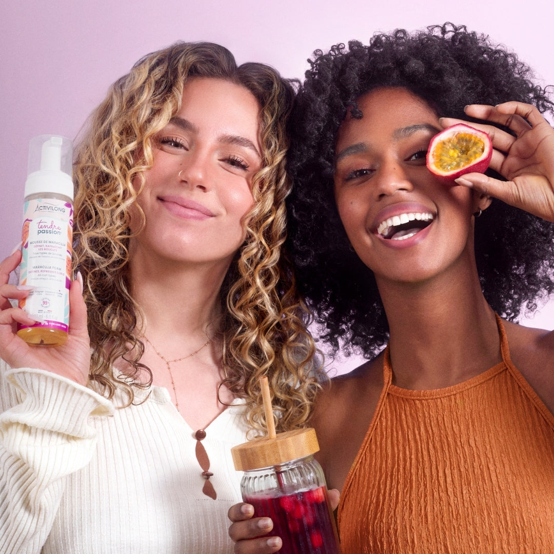 Deux femmes souriantes aux cheveux bouclés présentant le flacon-pousse-mousse de Mousse de maracuja Tendre Passion, l'une tenant un fruit de la passion coupé et une boisson, sur fond mauve.