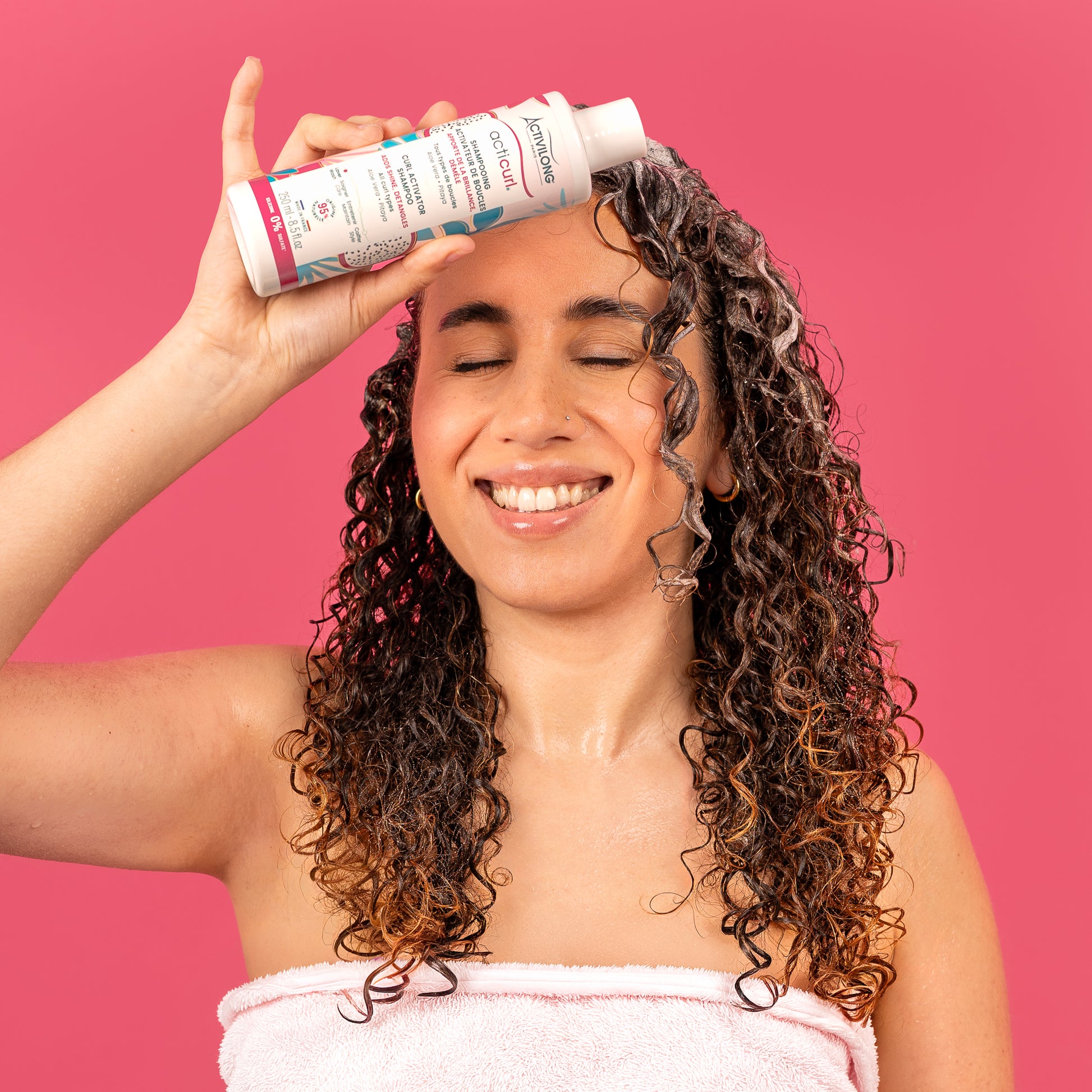 Visuel d'une femme aux cheveux bouclés humides, souriante, tenant le flacon Shampooing activateur de boucles Acticurl Activilong contre son front, sur fond rose vif
