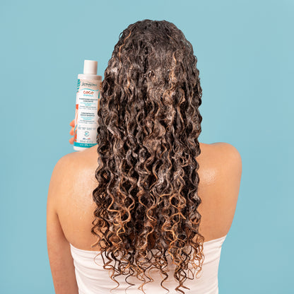 Femme de dos aux longs cheveux bouclés humides présentant le flacon de Shampooing douceur concentré Coco d'Amour, sur fond bleu.