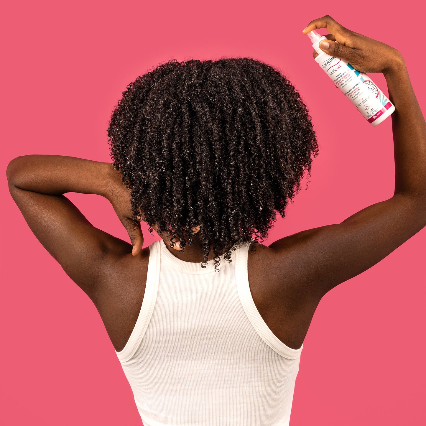 Visuel d'une femme vue de dos aux cheveux crépus volumineux vaporisant le Spray activateur de boucles Acticurl Activilong sur sa chevelure, sur fond rose vif