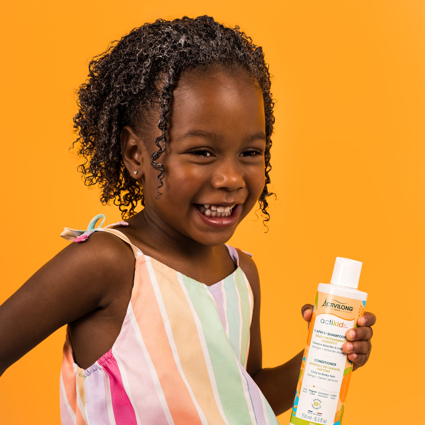 Visuel d'une enfant souriante aux cheveux frisés tenant le flacon du Ti Après-Shampooing Actikids sur fond orange