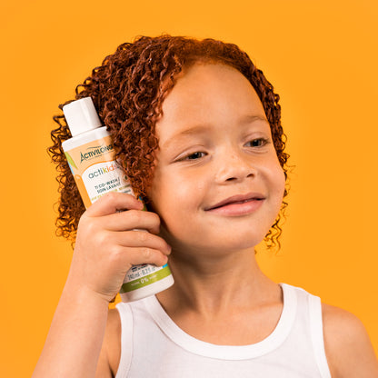 Visuel d'un enfant souriant aux cheveux bouclés roux tenant le flacon du Ti Co-Wash Actikids sur fond orange