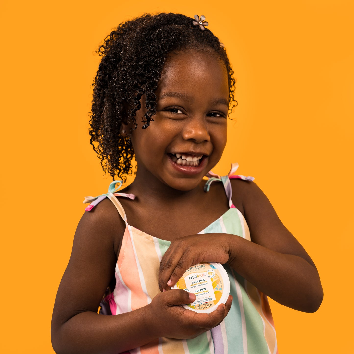 Visuel d'une enfant souriante aux cheveux bouclés tenant le pot du Hair Food Actikids sur fond orange