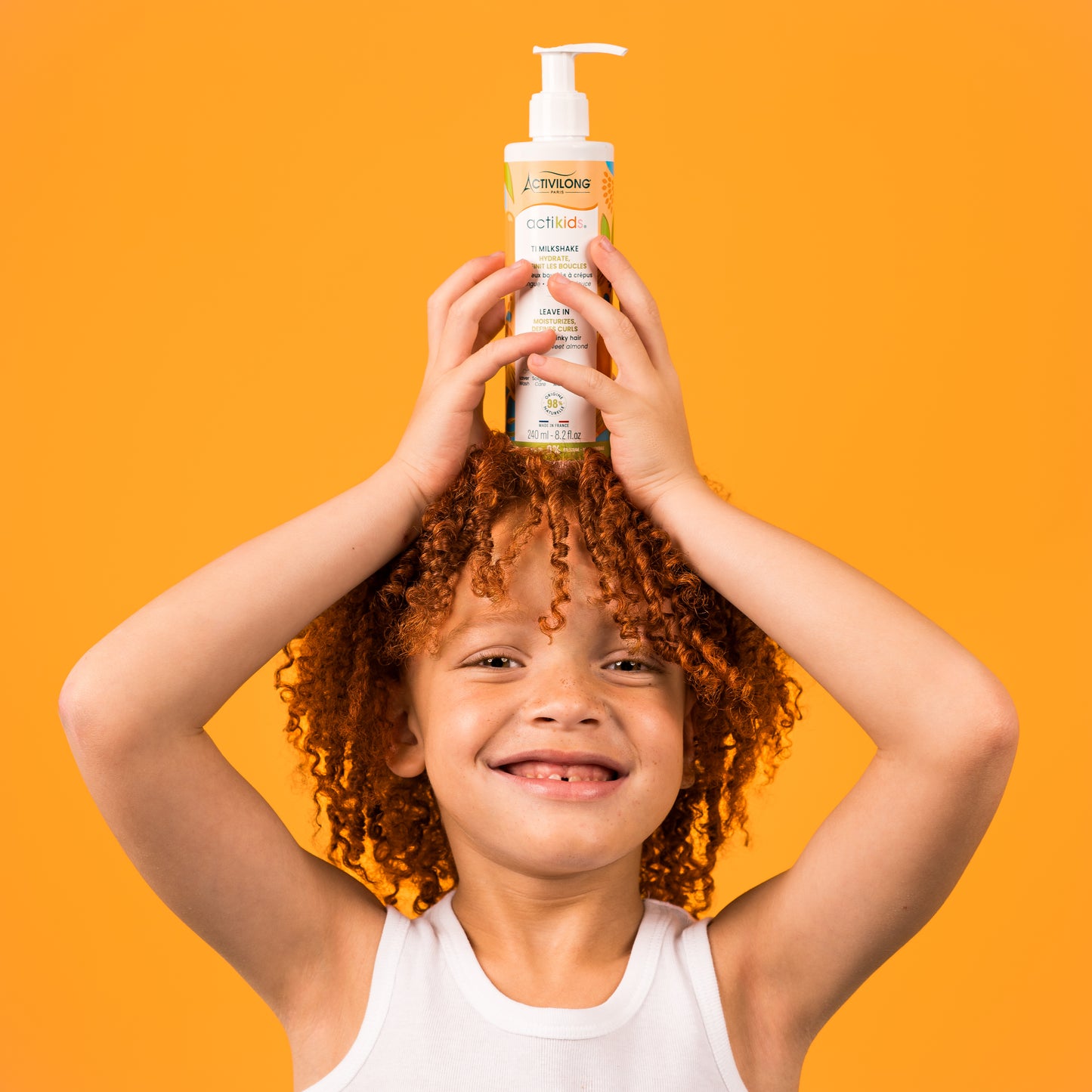 Enfant souriant aux cheveux bouclés roux tenant le flacon-pompe de Ti Milkshake / Leave-in Actikids au-dessus de sa tête, sur fond orange.