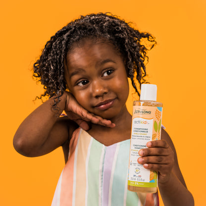 Petite fille souriante aux cheveux frisés présentant le flacon Ti Shampooing Conditionneur Actikids, sur fond orange.