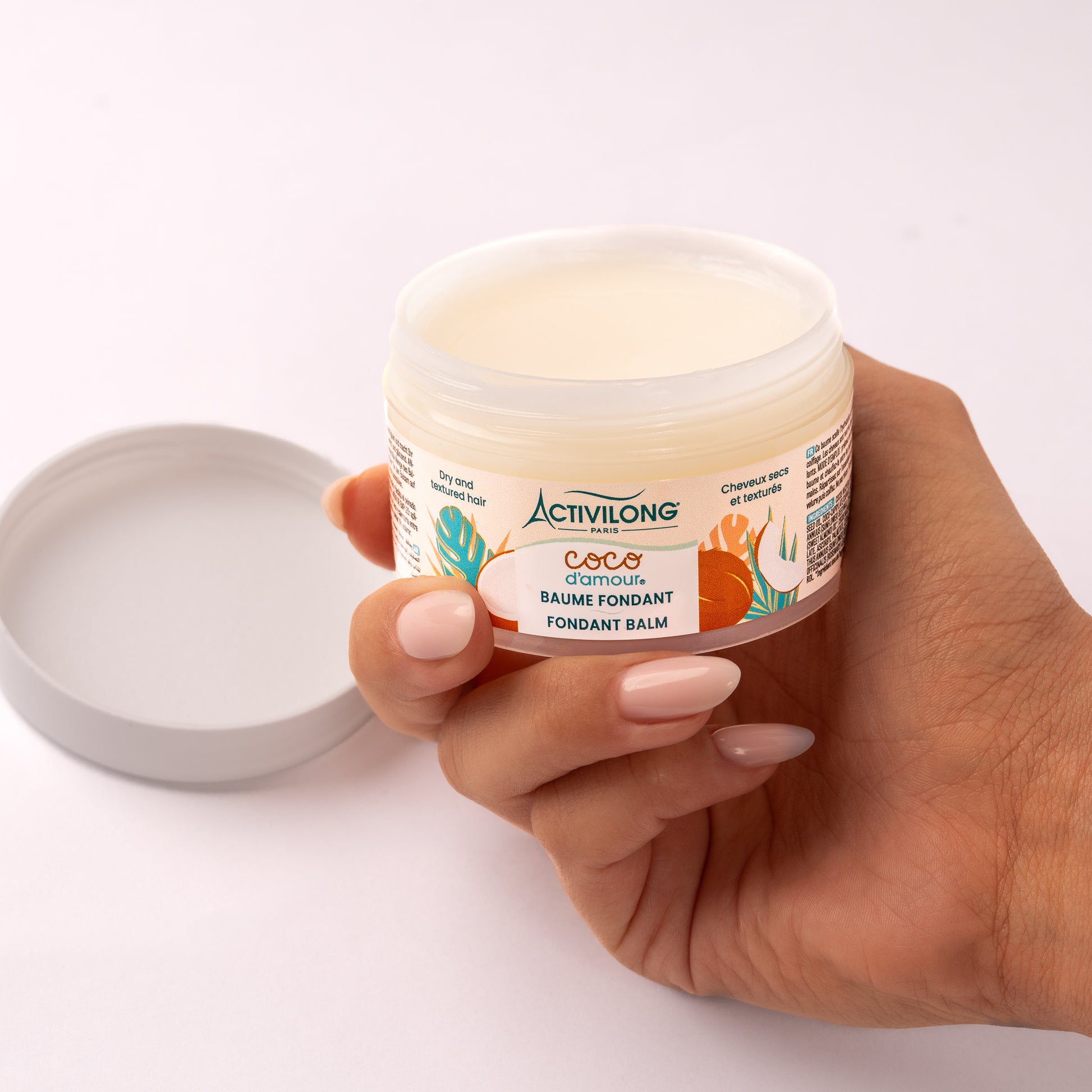 Pot de Baume fondant Coco d'Amour ouvert et tenu en main, couvercle posé à côté, révélant sa texture de baume riche blanc ivoire, sur fond gris clair.