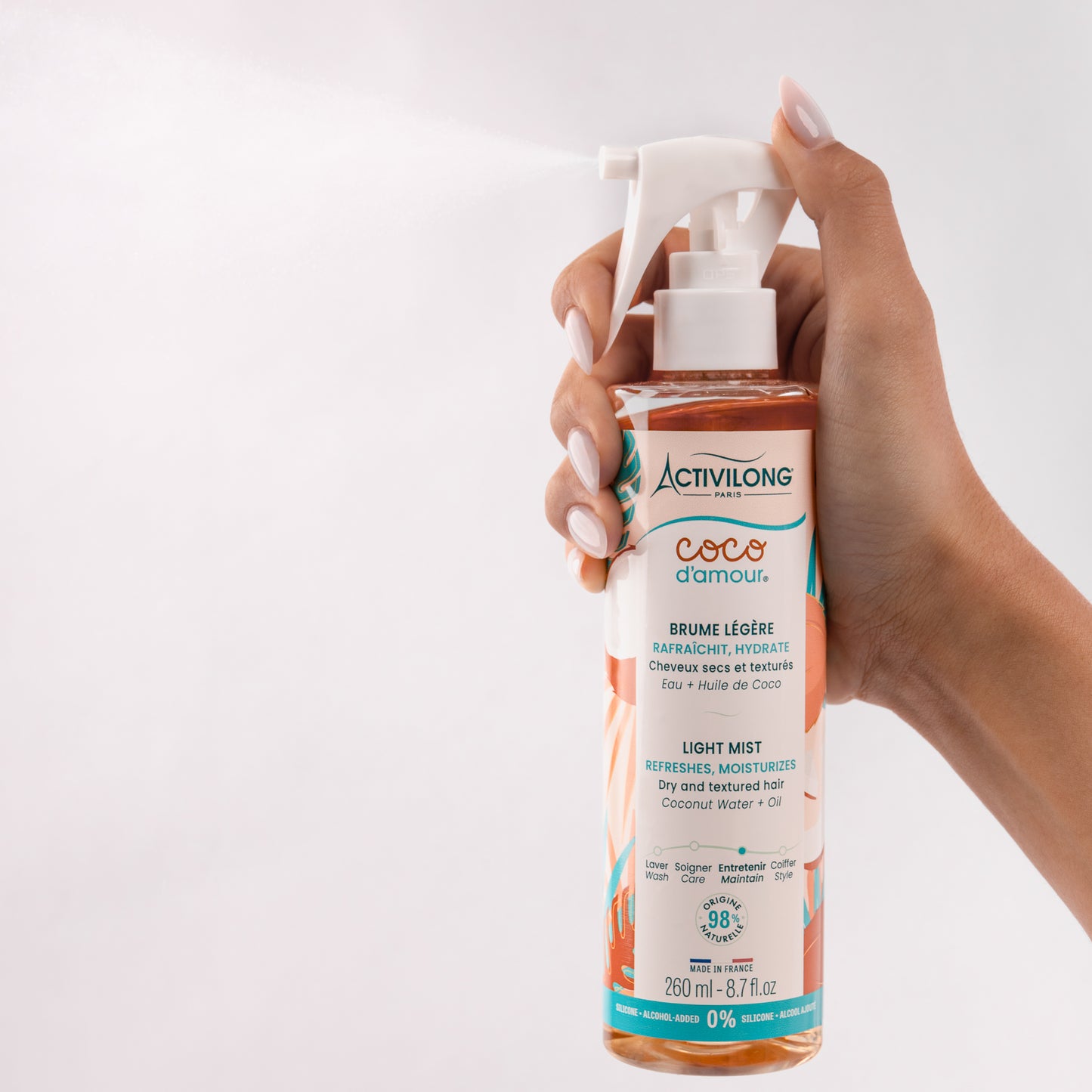 Flacon-spray de Brume légère Coco d'Amour tenu en main et actionné, diffusant un nuage de fines gouttelettes qui illustre sa texture vaporisée, sur fond gris clair.