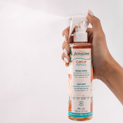 Flacon-spray de Brume légère Coco d'Amour tenu en main et actionné, diffusant un nuage de fines gouttelettes qui illustre sa texture vaporisée, sur fond gris clair.