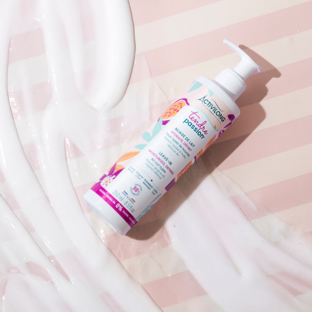 Visuel du flacon Nuage de lait Tendre Passion couché sur un fond rayé rose, entouré de la matière blanche du leave-in étalée
