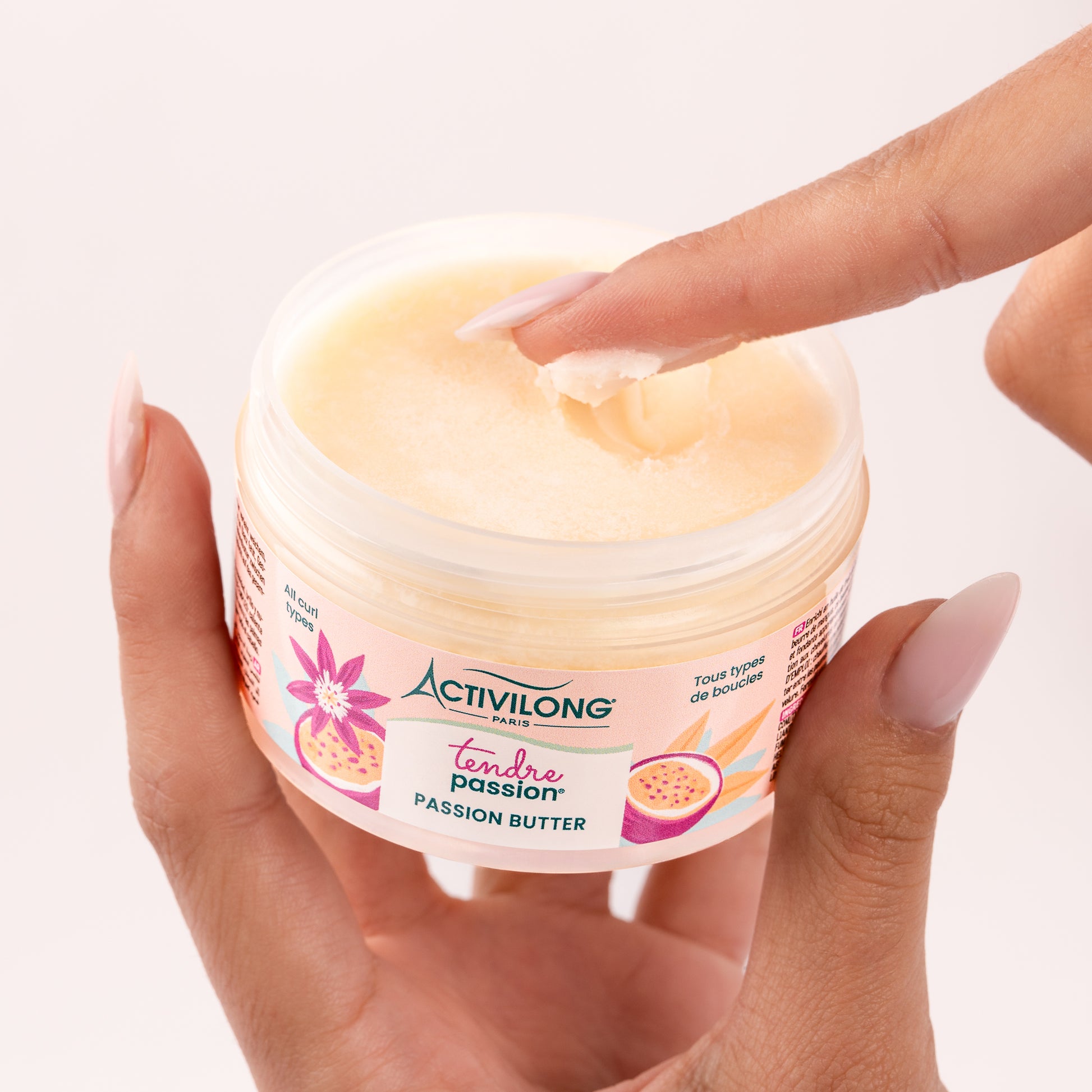 Pot ouvert de Passion butter Tendre Passion tenu en main, un doigt y prélevant une noisette de produit pour révéler sa texture de beurre riche jaune pâle, sur fond rose poudré clair.