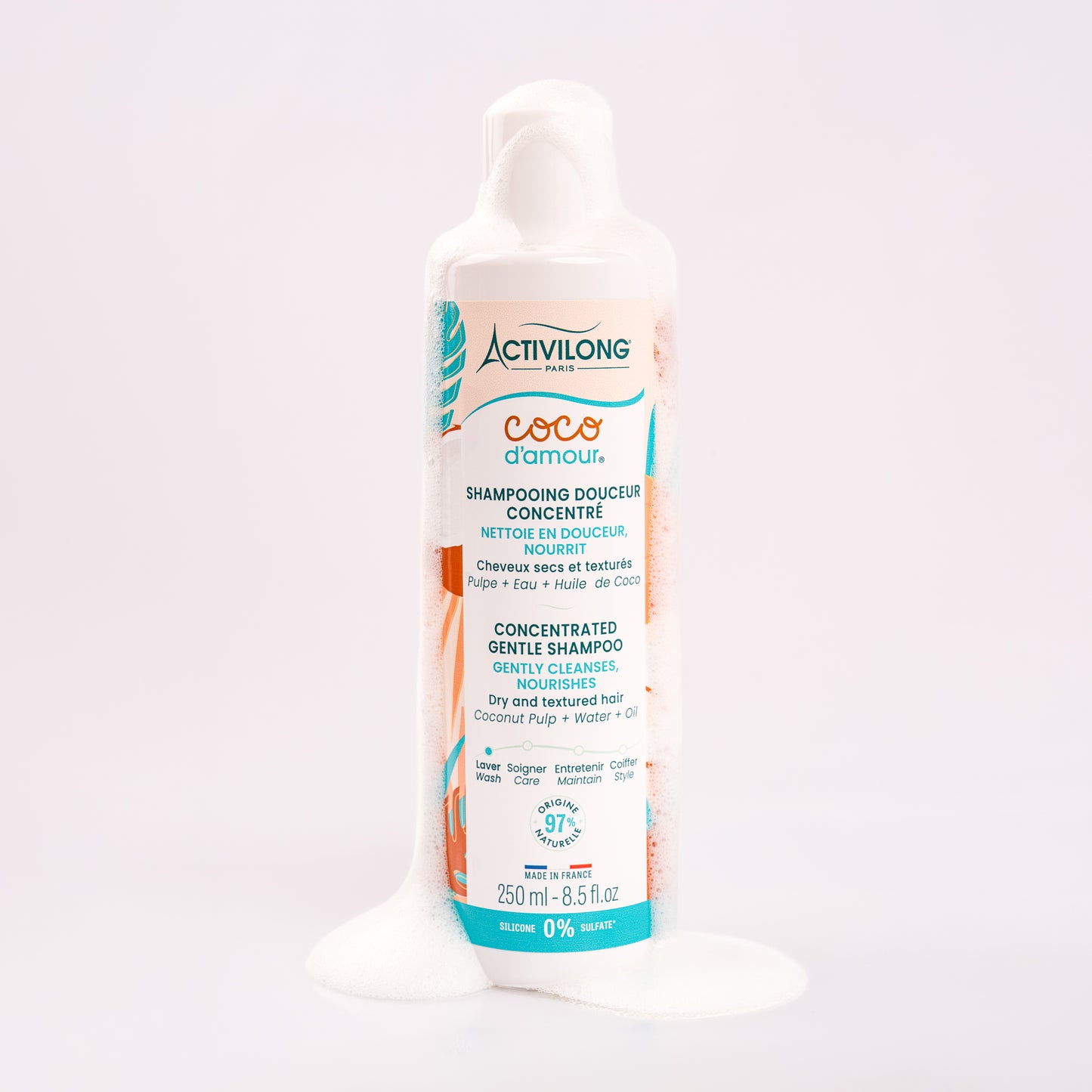 Flacon de Shampooing douceur concentré Coco d'Amour nappé de mousse onctueuse qui s'écoule le long du flacon et se répand à sa base, illustrant son pouvoir lavant moussant, sur fond gris clair rosé.