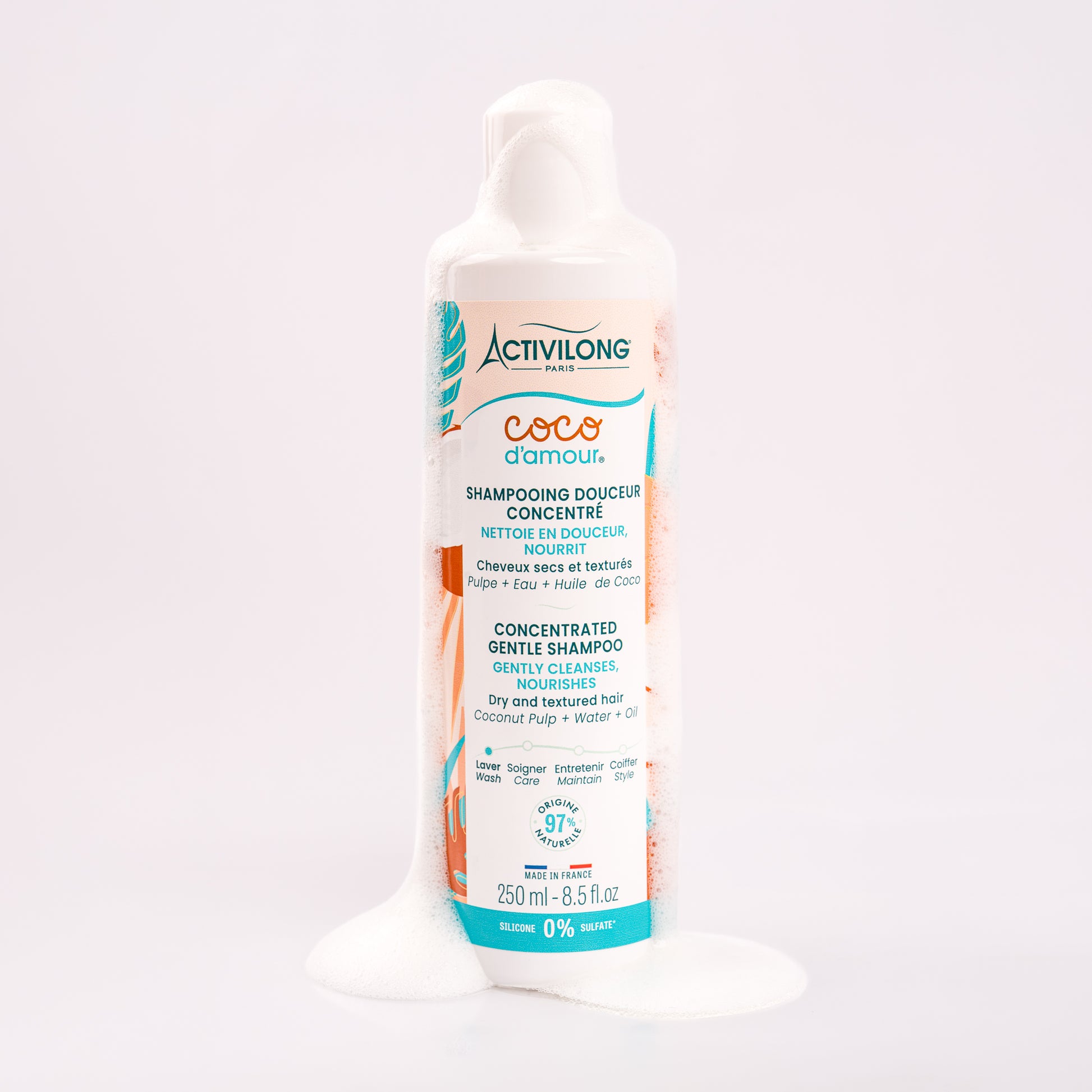 Flacon de Shampooing douceur concentré Coco d'Amour nappé de mousse onctueuse qui s'écoule le long du flacon et se répand à sa base, illustrant son pouvoir lavant moussant, sur fond gris clair rosé.