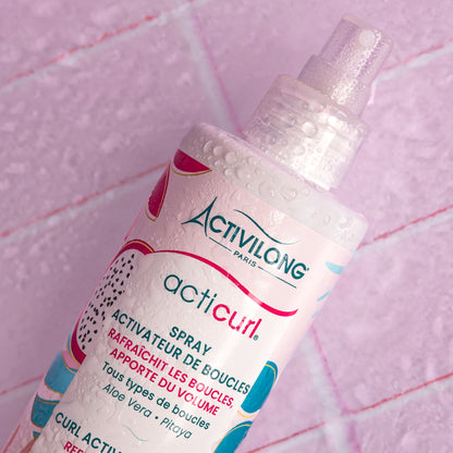 Visuel d'un Flacon du Spray Activateur de Boucles Acticurl en gros plan, couvert de gouttelettes d'eau sur fond rose