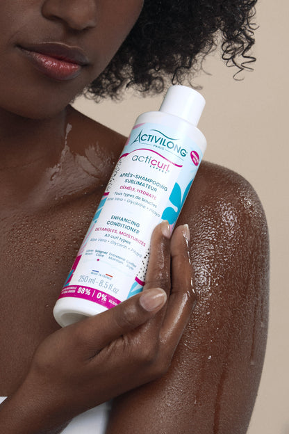 Modèle aux cheveux frisés tenant l’après-shampooing Sublimateur Acticurl Expert sur peau humide dans la douche