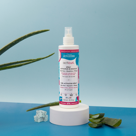 Spray Activateur de Boucles Acticurl posé sur un socle, avec feuilles d’aloe vera et morceaux de pitaya
